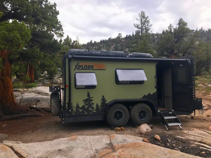 Off-road XPLORE RV rental parked in a rocky, forested landscape. Green trailer with open door, steps, and spare tire.. Imperial Outdoors XploreRV 2022