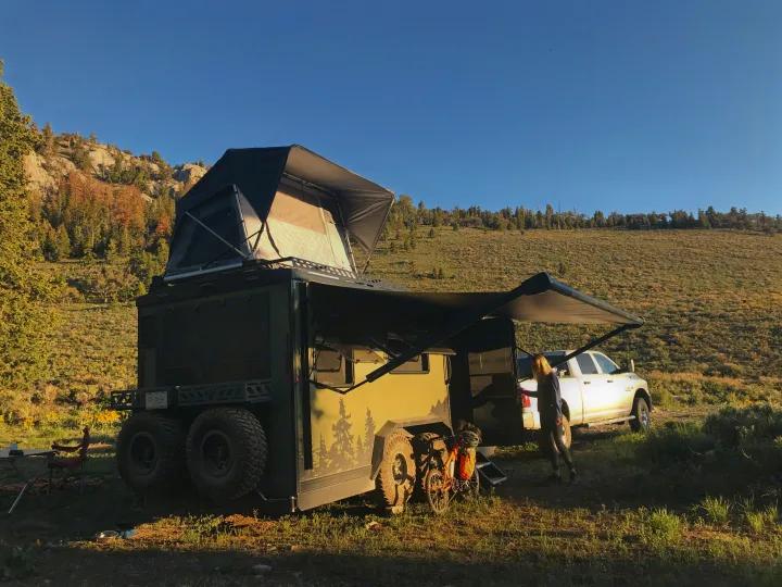 RV rental: Rugged overlanding trailer with rooftop tent & awning, pickup truck & person camping in scenic, hilly terrain.. Imperial Outdoors XploreRV 2022