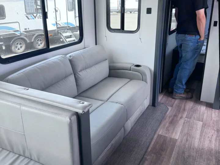 Experience an RV rental interior: grey reclinable sofa with cup holder, large windows, wood-look flooring. A person stands in the doorway.. Keystone RV Bullet Ultra Lite 2022