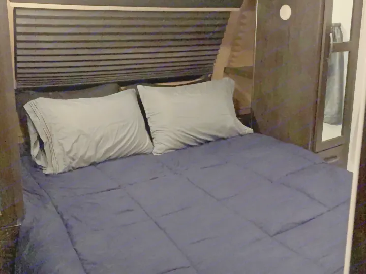 Dark RV rental bedroom with a bed, grey pillows, navy comforter, pleated blind, and built-in cabinet with mirror.. Forest River Surveyor Luxury 2019