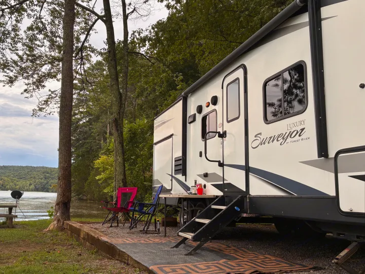This rv rental shows a "Surveyor" luxury travel trailer lakeside, with chairs & mat for a peaceful camping stay.. Forest River Surveyor Luxury 2019