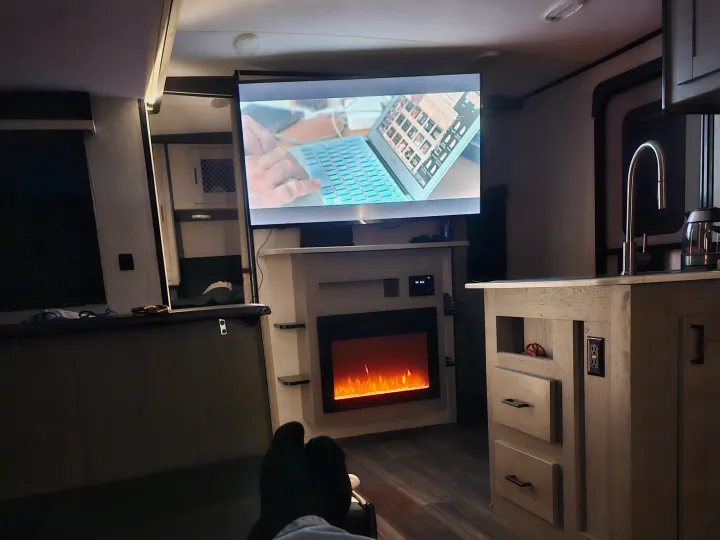 A person's socked feet rest in a cozy rv rental interior with TV, electric fireplace, and a small kitchen.. Heartland RVs Mallard 2021