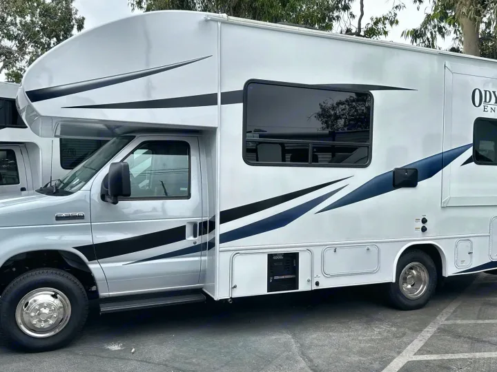 White RV rental parked outdoors, showcasing its side view with black and blue decals. Ideal for your next motorhome adventure.. Entegra Coach Odyssey SE 2025