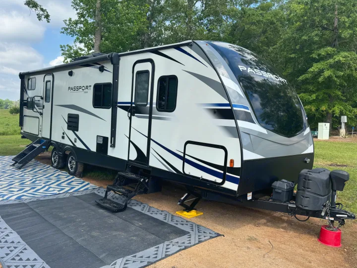 A modern Passport travel trailer RV rental, with steps, awning, and outdoor mats, parked in a scenic campground.. Keystone RV Passport Grand Touring 2019