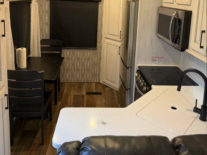 Modern RV rental interior: kitchen with white counters, black sink, stove, microwave, fridge; also a dining table & chairs.. Heartland RVs Bighorn Traveler 2022