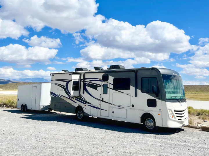 White Flair RV rental towing a trailer on a gravel lot under a bright, cloudy sky, ready for outdoor travel.. Flair-Fleetwood Flair-Fleetwood Motorhome 2020