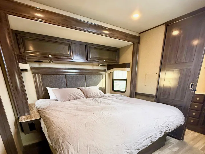 RV rental bedroom: large bed, dark wood headboard, overhead cabinets, and window. A cozy, well-lit interior for travel.. Flair-Fleetwood Flair-Fleetwood Motorhome 2020