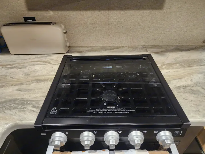 RV rental kitchen: black Furrion stove with a clear glass cover, and a light-colored Braun toaster on the counter.. Grand Design Transcend Xplor 2022