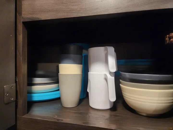 RV rental kitchen cabinet showing stacked durable cups, plates, and bowls in black, grey, cream, and blue colors.. Grand Design Transcend Xplor 2022