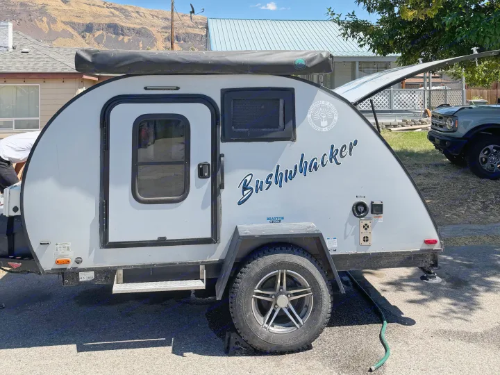 Grey Bushwhacker teardrop RV rental parked, ready for an adventure trip, with a house and mountain backdrop.. Braxton Creek Bushwhacker 2022