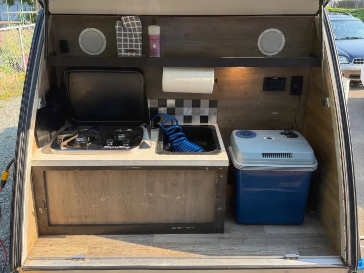Compact RV rental kitchen with stove, sink, cooler, and storage in an open camper hatch, ready for use.. Braxton Creek Bushwhacker 2022