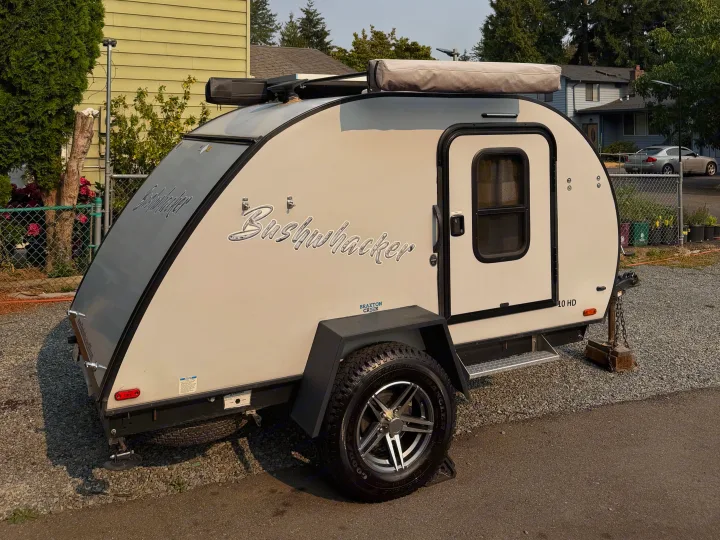 Beige & gray Bushwhacker teardrop camper, ideal for rv rental. This compact travel trailer with an awning is parked on gravel.. Braxton Creek Bushwhacker 2022