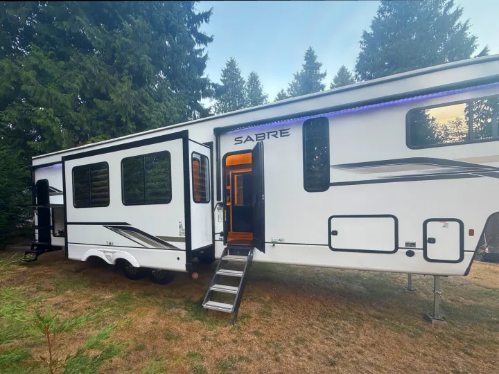 White Sabre RV rental parked with door open, steps out, & blue awning lights on, nestled in a grassy campsite with trees.. Forest River Sabre 2025