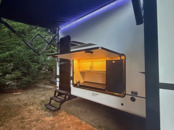 Evening view of an RV rental's exterior, featuring an illuminated outdoor kitchen, awning with LED lights, and steps. Ideal for camping.. Forest River Sabre 2025