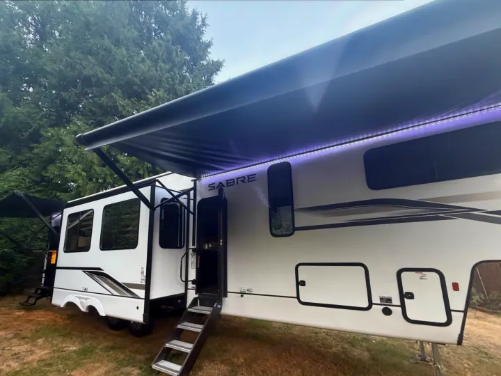White Sabre fifth-wheel RV rental with steps and illuminated awning, set up outdoors for a camping getaway.. Forest River Sabre 2025