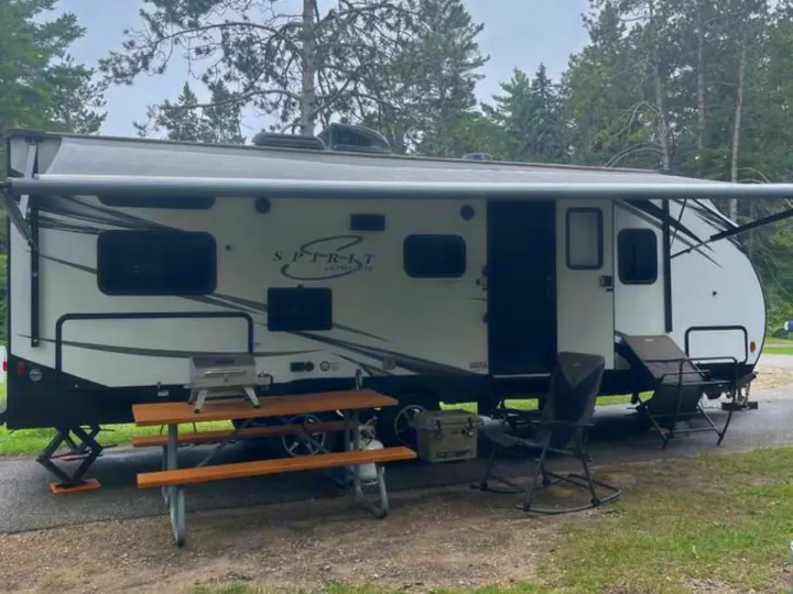 Relaxing rv rental campsite: A white travel trailer with an awning, picnic table, grill, cooler, and chairs in a wooded area.. Coachmen Spirit 2019
