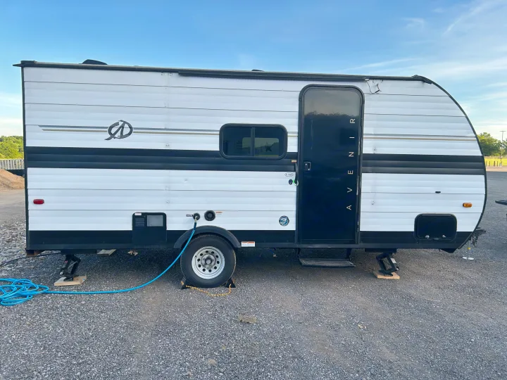 White & black travel trailer rv rental on gravel. Blue hose connected near wheel, leveling jacks visible. Ready for camping.. Cruiser RV A-17BH Avenir 2024