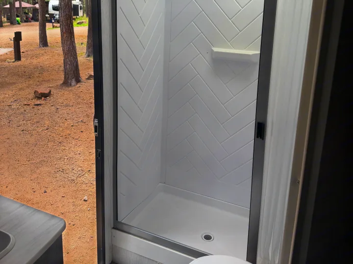 Modern RV rental shower stall with herringbone walls, offering a view of a rustic campsite with trees and another camper.. Forest River Salem Cruise Lite 2022
