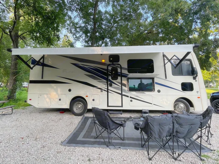 This RV rental offers outdoor entertainment with an extended awning, external TV, and comfy chairs on a gravel campsite.. Flair-Fleetwood Flair-Fleetwood Motorhome 2022