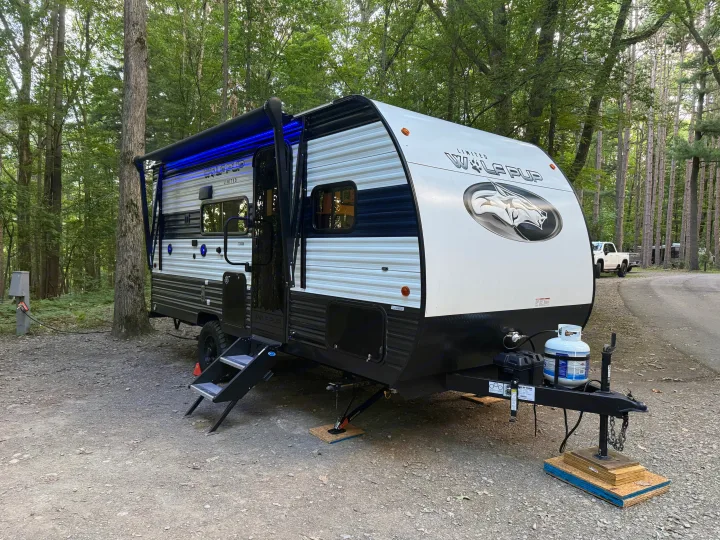 Wolf Pup rv rental travel trailer with blue awning lights, parked at a wooded campsite on a gravel pad.. Forest River Cherokee Wolf Pup 2024