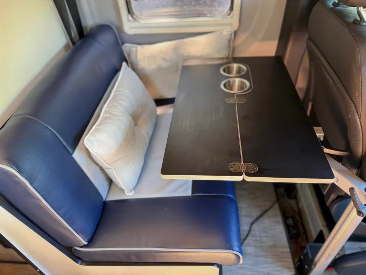 Cozy RV rental interior showing a blue upholstered bench, light pillows, and a black table with built-in cup holders.. Winnebago Revel 2022