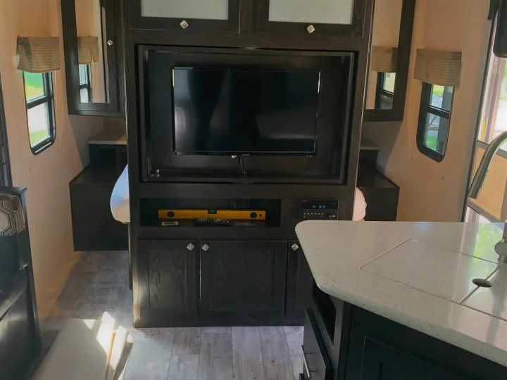 RV rental interior showing a dark wood entertainment center with TV, a level, remotes, and a light speckled kitchen counter with sink.. Heartland RVs North Trail 2019
