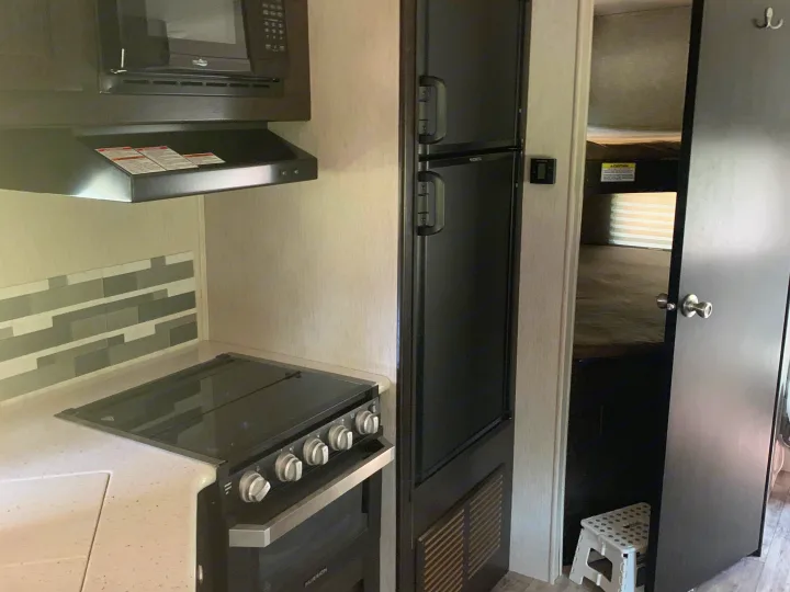 This RV rental shows a compact kitchen with a microwave, stove, refrigerator, and a doorway leading to bunk beds.. Heartland RVs North Trail 2019