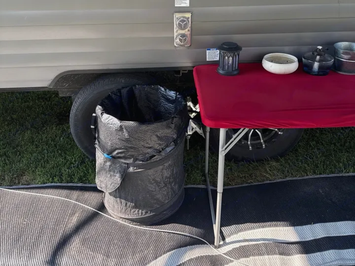RV rental outdoor setup: A red table with camping items, a pop-up trash can, and a mat by the camper tire.. Forest River Campsite Reserve 2025
