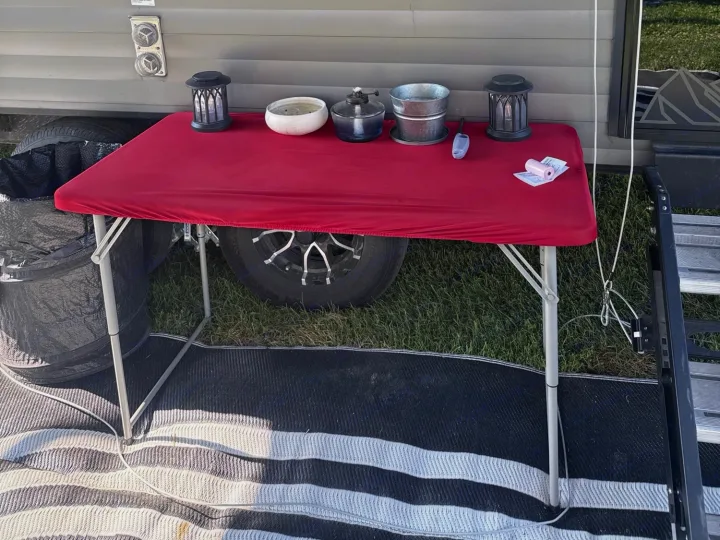 RV rental outdoor setup: A red folding table with lanterns, bowls, and pet waste bags, on a striped mat next to the camper.. Forest River Campsite Reserve 2025