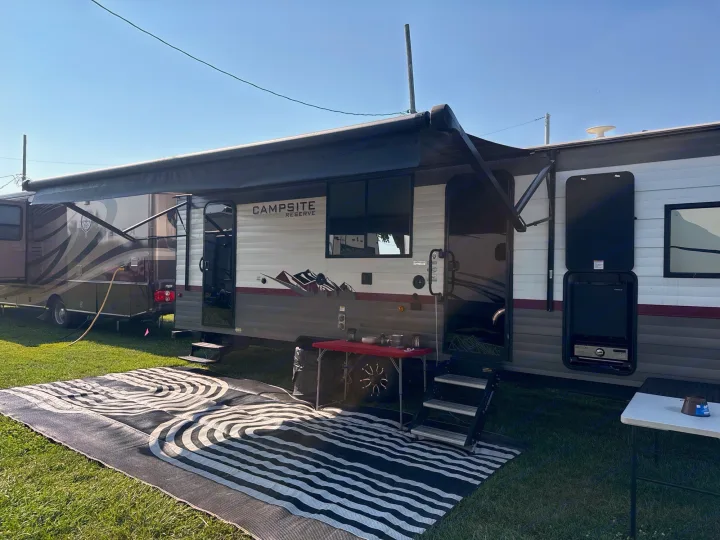 An RV rental with an extended awning, outdoor rug, and table on a sunny, grassy campsite.. Forest River Campsite Reserve 2025