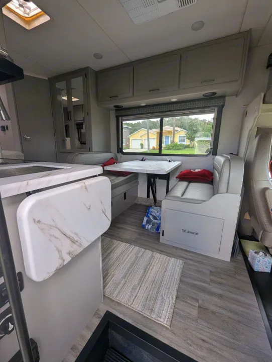 RV rental interior showcasing a contemporary dinette with seating, kitchen counter, overhead cabinets, and a bright window.. Thor Quantum SE 2025