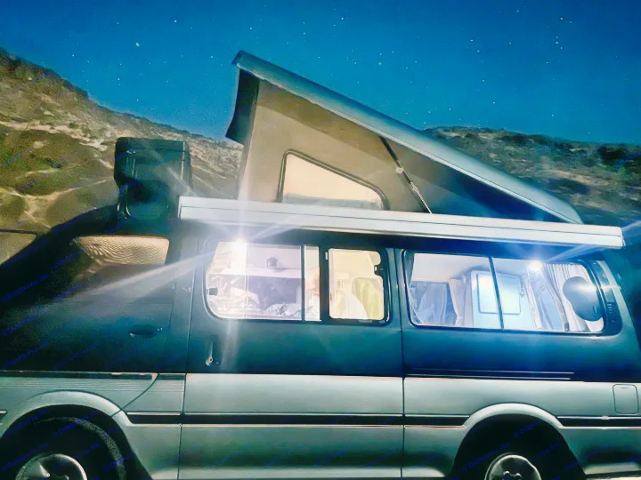 Cozy rv rental camper van under a starry night sky, with its pop-top extended and interior lights glowing, parked outdoors.. Toyota Hiace Campervan 1994