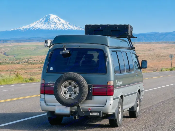 Adventure awaits! This RV rental van with rooftop storage is parked on a scenic road, with a snow-capped mountain backdrop.. Toyota Hiace Campervan 1994