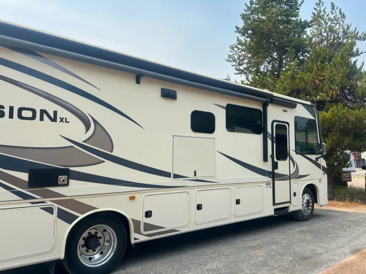 This cream-colored RV rental, with dark stripes, is parked on gravel by tall trees, ready for adventure.. Entegra Coach Vision XL 2021