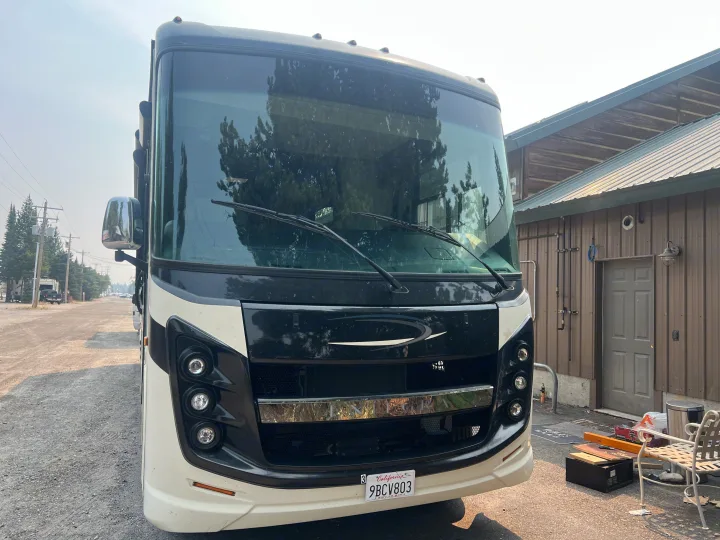 Front of a sleek, black and white RV rental parked on a gravel lot. Reflections of trees are visible on its large windshield.. Entegra Coach Vision XL 2021