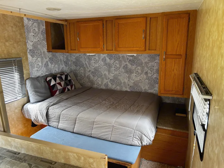 This cozy rv rental bedroom features a bed with gray bedding, floral wallpaper, and wood storage cabinets above. A compact space for travel.. Coachmen Spirit of America 2009