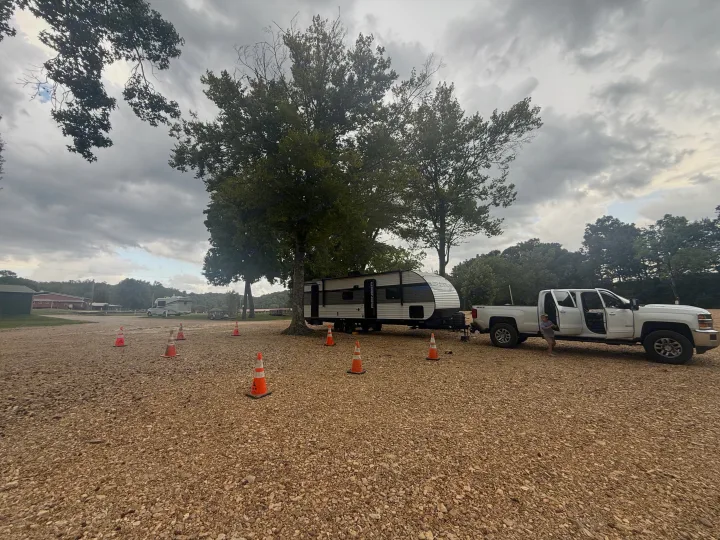 A white pickup tows an RV rental in a gravel campground, marked by orange cones. A child is near the truck. Cloudy day scene.. Heartland RVs Pioneer 2024