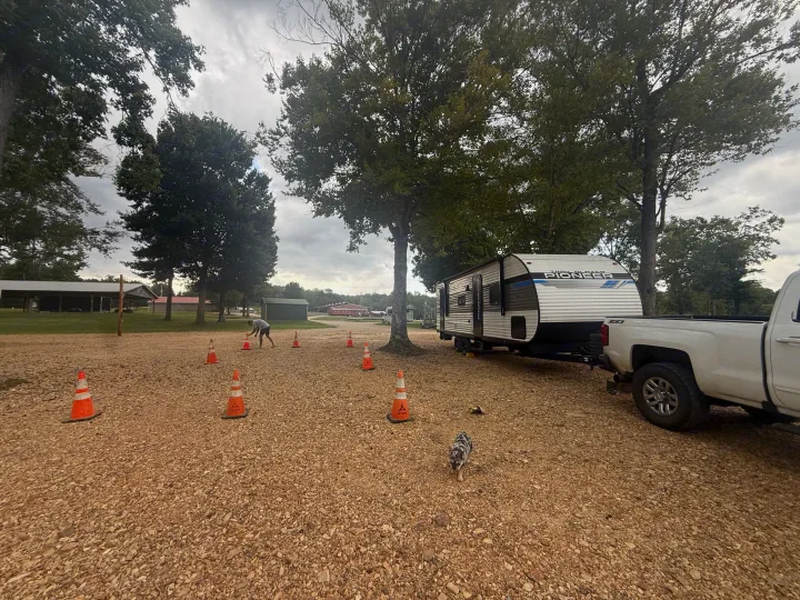 RV rental hitched to a pickup truck, parked on a gravel site marked by orange cones, with a person and dog.. Heartland RVs Pioneer 2024