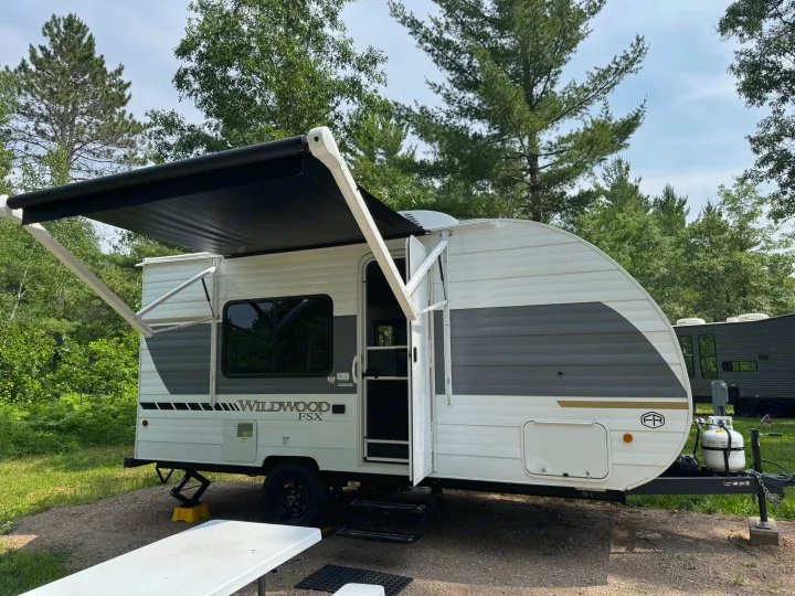 This white and gray Wildwood FSX travel trailer, an ideal rv rental, is parked with its awning extended at a serene campsite.. Forest River Wildwood FSX 2026