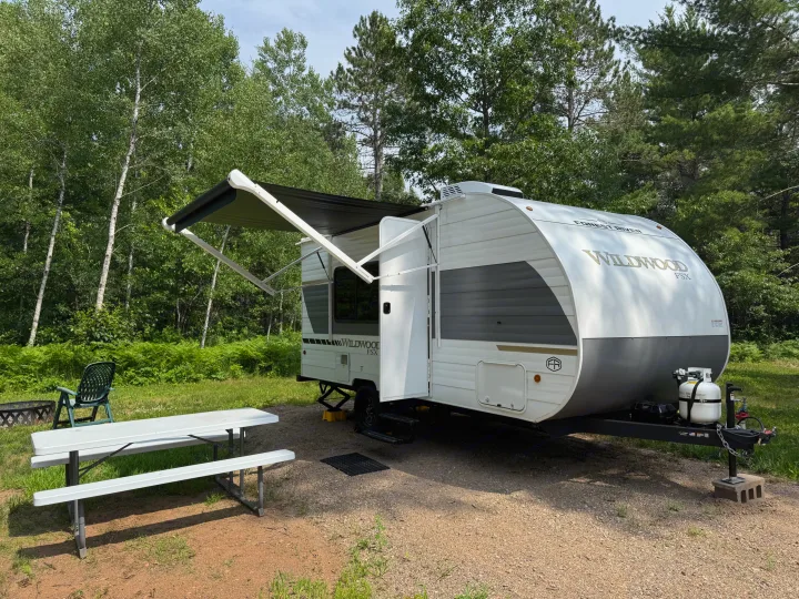 White & gray RV rental travel trailer with awning extended, parked on a gravel campsite with picnic table, chair & fire pit.. Forest River Wildwood FSX 2026