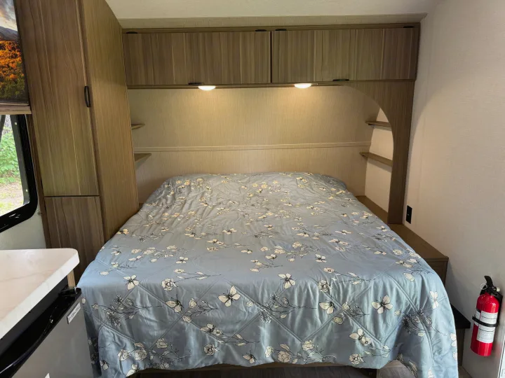 This RV rental bedroom features a comfy bed with a floral comforter, storage cabinets, and a safety fire extinguisher.. Forest River Wildwood FSX 2026