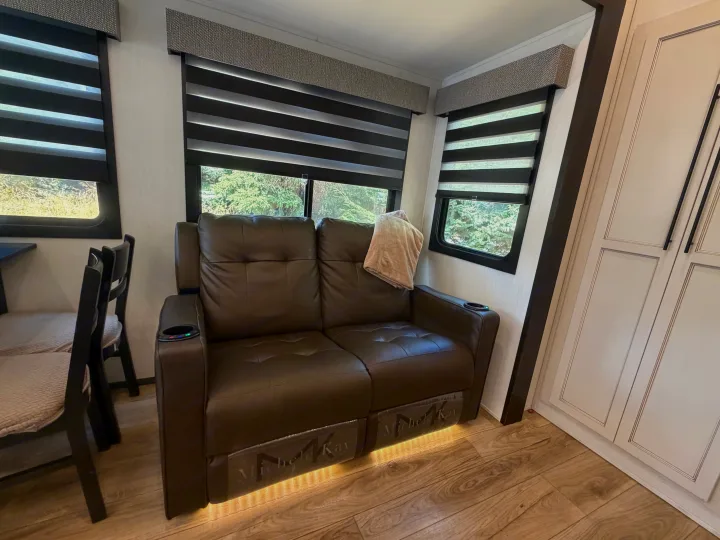 Cozy RV rental living space with a brown leather reclining loveseat, illuminated base, cup holders, and windows with blinds.. Forest River Sabre 2025