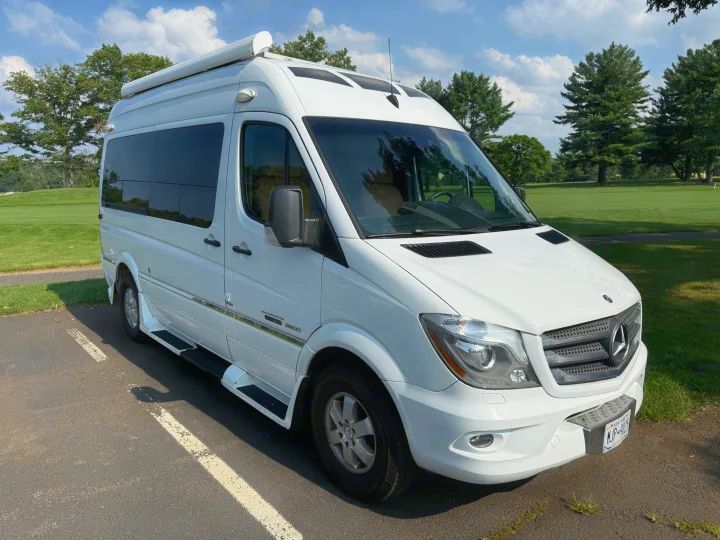 White Mercedes-Benz Sprinter rv rental van parked on asphalt. A green golf course & trees are visible under a blue sky.. Roadtrek Roadtrek Class B 2014