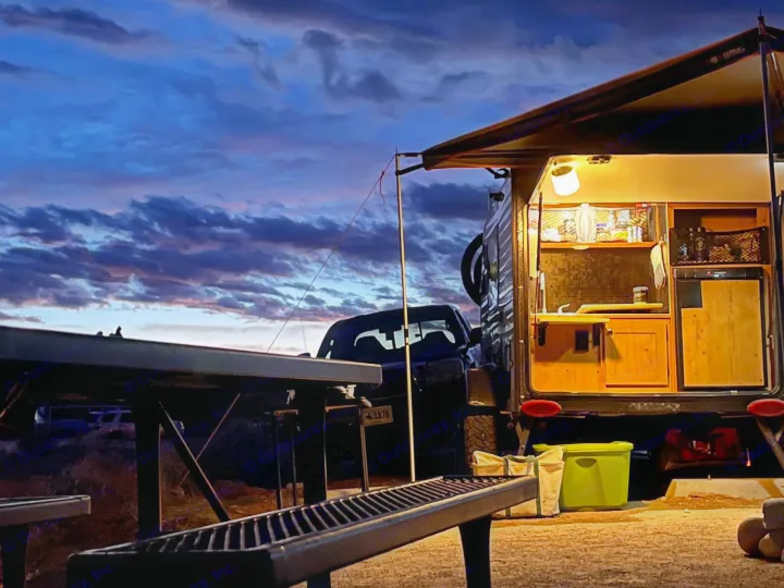 Evening glow on a compact RV rental's illuminated kitchen setup under a dramatic twilight sky. Truck & picnic table visible for camping.. Sunset Park RV SunRay 2020
