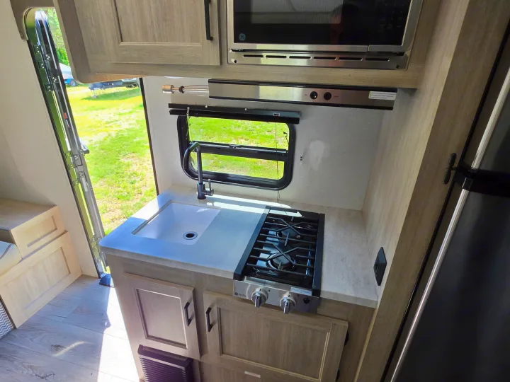 Compact kitchen in an rv rental, featuring a sink, stovetop, and microwave, with an open door leading to the outdoors.. Coachmen 184BHS 2024