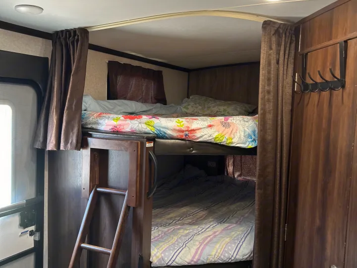 Inside an RV rental: two bunk beds with a ladder, floral & striped bedding, privacy curtains, & a coat rack.. Jayco White Hawk 2019