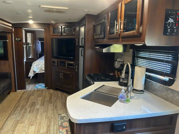 Spacious rv rental interior showing a kitchen, living area, and bedroom, with dark wood cabinets and light countertops.. Jayco White Hawk 2019