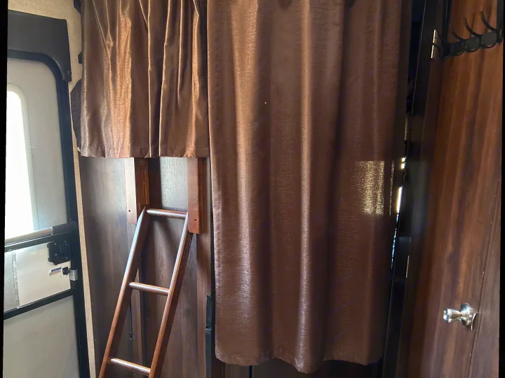 RV rental interior with a wooden bunk bed ladder, brown privacy curtains, a door with a silver knob, and a coat rack.. Jayco White Hawk 2019