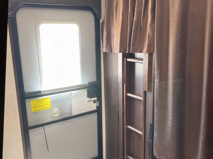 RV rental interior: bright entry door, screen, and dark curtains partly open revealing a bunk bed ladder.. Jayco White Hawk 2019