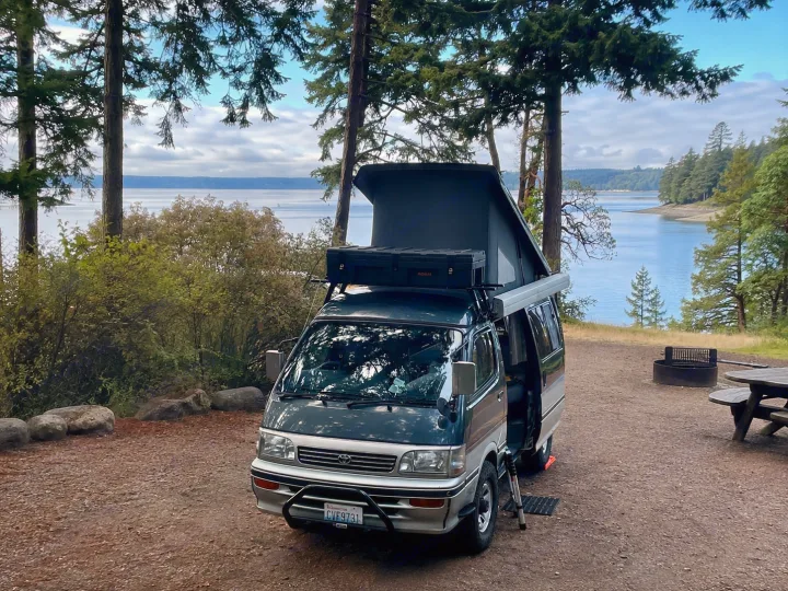 This dark green pop-top camper van, ideal for RV rental, is parked by a scenic lake campsite with tall trees.. Toyota Hiace Campervan 1994